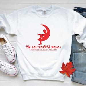 May include: White sweatshirt with a red graphic of a person sitting on a crescent moon. The text "ScreamWorks Never Sleep Again" is printed below the graphic.