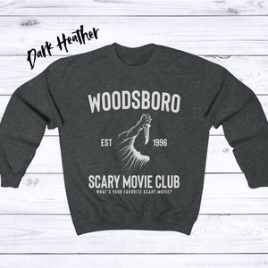 May include: Dark heather grey sweatshirt with a white graphic design. The graphic features a hand holding a knife with the text "Woodsboro Est 1996 Scary Movie Club What's your favorite scary movie?"