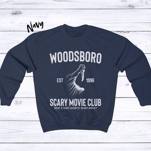 May include: Navy blue sweatshirt with a white graphic design that says "Woodsboro Est. 1996 Scary Movie Club What's your favorite scary movie?"