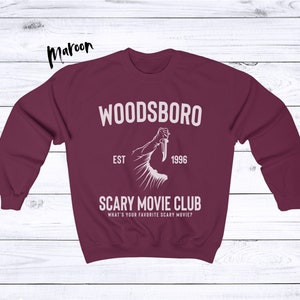 May include: Maroon sweatshirt with white text that reads "Woodsboro Est. 1996 Scary Movie Club What's your favorite scary movie?" and a graphic of a hand holding a knife.