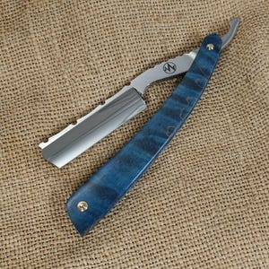Könnte beinhalten: Ein Rasiermesser mit einer polierten, silberfarbenen Metallklinge und einem Griff aus blauem Holz mit sichtbarer Maserung. Die Metallangel zeigt ein kreisförmiges Logo mit den Buchstaben AN. Ein kleiner goldfarbener Niet sichert den Griff.