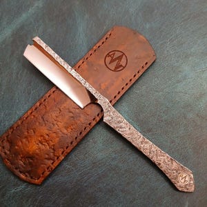 Könnte beinhalten: Ein Rasiermesser mit einer polierten Metallklinge und einem strukturierten, dunkelgrauen, gehämmerten Metallgriff mit einem kleinen kreisförmigen Logo am Ende, das auf einer braunen Lederscheide mit passendem Logo liegt.