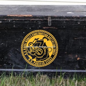 May include: A weathered, black wooden chest with a metal clasp. A yellow and black emblem is affixed to the front, displaying the text "U.S. Naval Mobile Construction Battalion THREE U.S. PACIFIC FLEET". The chest is set on grass.