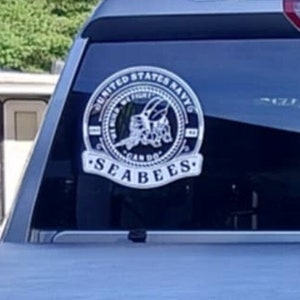 May include: A silver and black sticker on a car window. The sticker features a circular design with the text "United States Navy" and "Seabees" around the edge. A stylized bee with wings is in the center of the circle.
