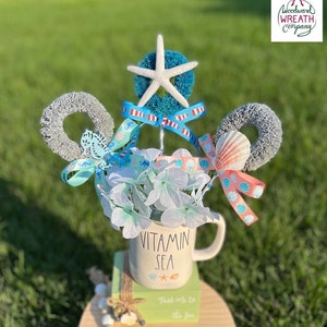 May include: A summer-themed centerpiece featuring a white mug with the text "Vitamin Sea" and a starfish on a blue and white background. The centerpiece also includes a blue and white wreath, a seashell, and a white sand dollar. The centerpiece is on a wooden tiered stand with a blue and white fringe.