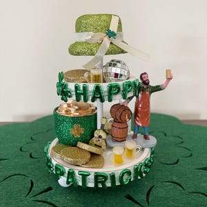 May include: A white tiered stand decorated for St. Patrick's Day. The top tier has a green glitter top hat with a white ribbon and a disco ball. The middle tier has a green glitter jar with gold coins, a wooden barrel, and a miniature figure of a man holding a beer. The bottom tier has gold coins, two miniature beer mugs, and the words "Happy St. Patrick's Day" in green letters.