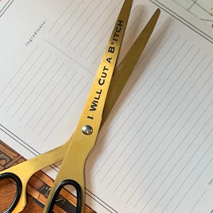 May include: Gold-colored scissors with black handles, resting on a white lined paper. The scissors have the text "I WILL CUT A B*TCH" printed on one blade. The scissors are likely for crafting or general use.