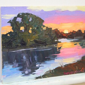 May include: Oil painting of a sunset over a river. The sky transitions from purple to orange and pink, reflecting in the water. Dark green trees line the riverbank. The artwork is on a small canvas.