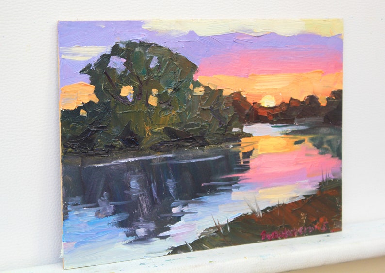 May include: Oil painting of a sunset over a river. The sky transitions from purple to orange and pink, reflecting in the water. Dark green trees line the riverbank. The artwork is on a small canvas.