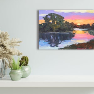 May include: An original oil painting depicts a sunset over a calm river. The sky transitions from pink to orange and purple, reflecting in the water. Dark green trees line the riverbanks, creating a serene landscape scene.