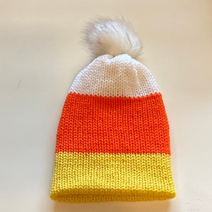 Op de afbeelding: Een gebreide muts in de stijl van snoepmaïs. De muts heeft horizontale strepen in wit, oranje en geel. Bovenop de muts is een pluizige, witte pompon bevestigd. Perfect voor Halloween of de herfst.