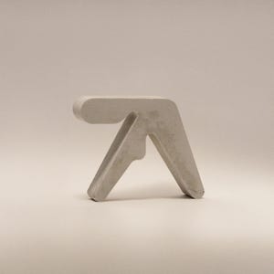 Może przedstawiać: Mały, jasnoszary betonowy stołek o minimalistycznym designie. Stołek ma zakrzywioną górę i dwie skośne nogi, tworząc nowoczesny, geometryczny kształt. Ogólna estetyka jest prosta i industrialna.