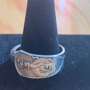 Può includere: Un anello d'argento con un designo dettagliato di una figura che tiene una palla. L'anello è su un espositore in velluto nero.
