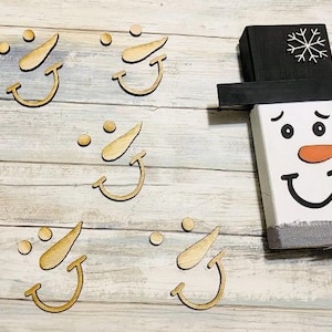 May include: A wooden snowman with a black hat and a carrot nose. The snowman has a gray scarf and a smiling face. Scattered around are wooden snowman features: smiles, carrot noses, and dots.