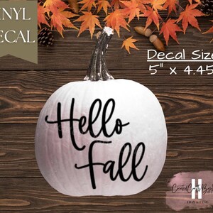 May include: A white pumpkin featuring a black vinyl decal with the text Hello fall in a cursive script font. The decal measures 5 inches by 4.45 inches. The pumpkin has a dark brown stem and is displayed against a dark wood background with leaves.