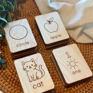 Könnte beinhalten: Holz-Lernkarten mit Illustrationen und Wörtern. Die Karten zeigen einen Kreis, einen Apfel, eine Katze und die Zahl eins mit einer Sonne. Die Karten sind auf einem gewebten Tablett gestapelt.