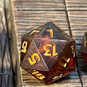 May include: A translucent amber-colored twenty-sided die with gold numbers. The die is sitting on a wooden surface.
