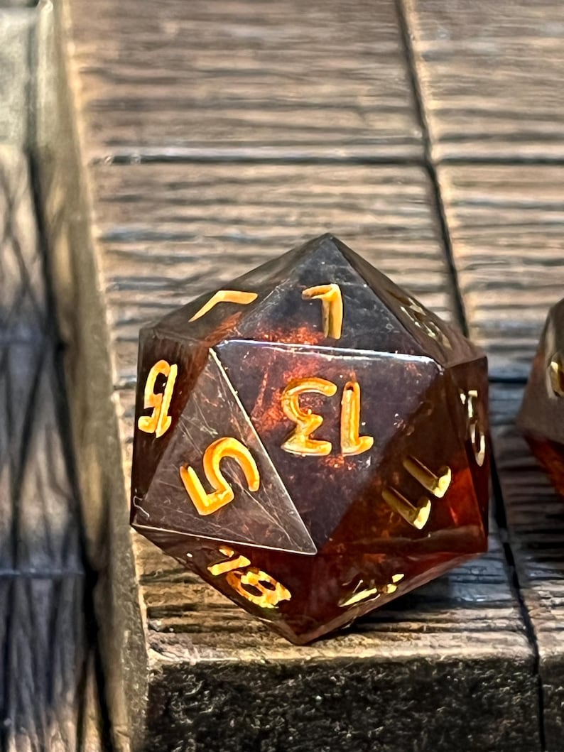 May include: A translucent amber-colored twenty-sided die with gold numbers. The die is sitting on a wooden surface.