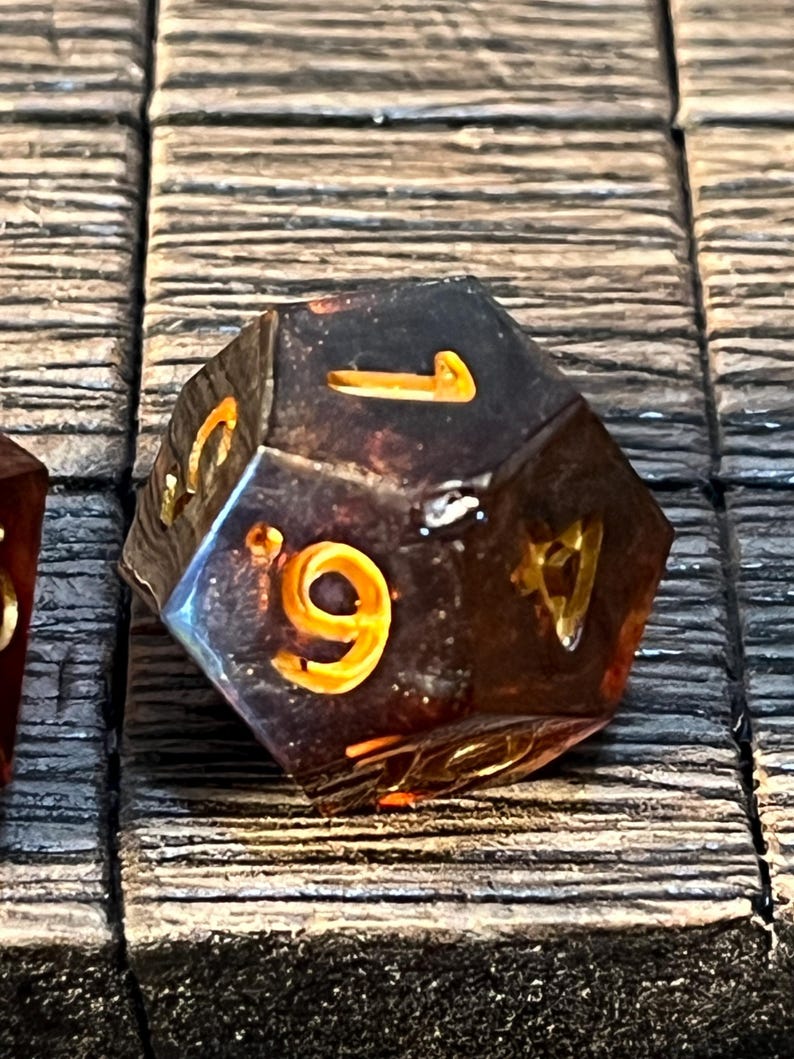 May include: A dark amber-colored, multi-sided dice with gold numbers. The dice is sitting on a wooden surface.