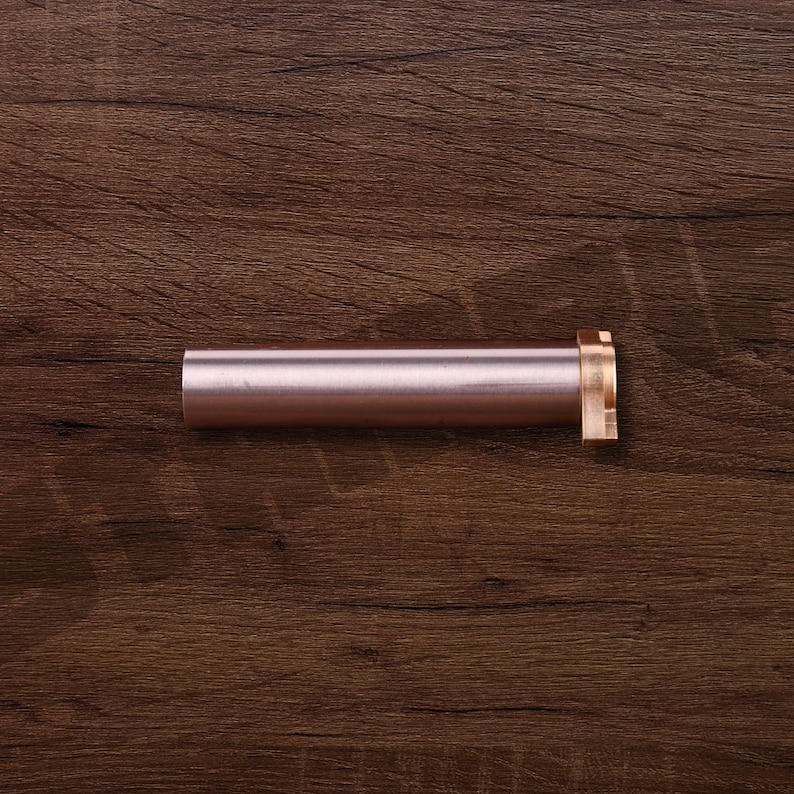 K&ouml;nnte beinhalten: Ein zylindrisches Metallobjekt in Ros&eacute;gold mit einem messingfarbenen Flansch an einem Ende. Das Objekt liegt horizontal auf einer dunkelbraunen Holzoberfl&auml;che. Das Objekt scheint eine Komponente oder ein Teil eines gr&ouml;&szlig;eren Artikels zu sein.