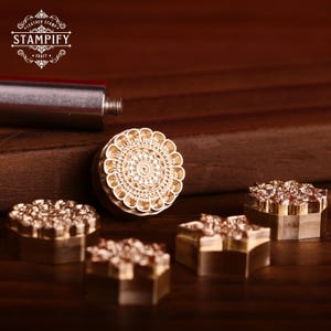 Puede incluir: Primer plano de varios sellos de cuero de latón con diseños florales y geométricos. Los sellos, incluyendo una flor y una estrella, se muestran sobre una superficie de madera. El logotipo "STAMPIFY" es visible. Herramientas para artesanía en cuero.