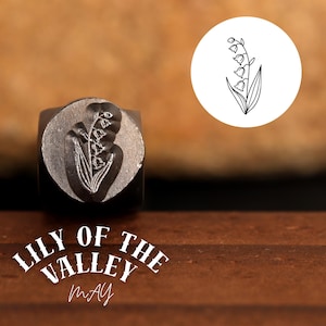 Może przedstawiać: Metalowa stempla z wzorem kwiatu konwalii. Stempla leży na drewnianej powierzchni z tekstem "Lily of the Valley May" poniżej.