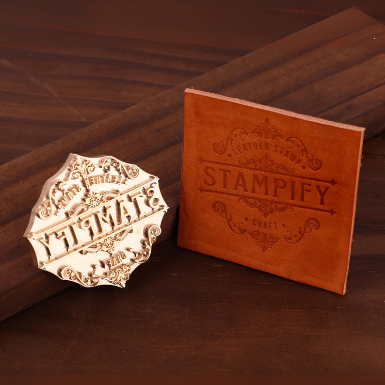 K&ouml;nnte beinhalten: Ein Holzstempel mit dem Wort "ULTIMATE" in einer dekorativen Schriftart und ein Lederquadrat mit dem Wort "STAMPIFY" darauf gestempelt. Der Stempel und das Leder liegen auf einer Holzoberfl&auml;che.