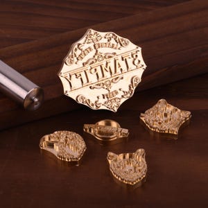 K&ouml;nnte beinhalten: Eine Sammlung von Messingstempeln mit aufwendigen Designs, darunter einer mit dem Wort "STAMPATE" in einem dekorativen Rahmen. Links ist ein silberner Metallgriff zu sehen. Die Stempel dienen zum Basteln und Erstellen individueller Designs.