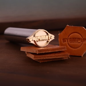 K&ouml;nnte beinhalten: Ein Metallstempel mit dem Text "STAMPIF" und "METAL STAMP" auf einem Lederst&uuml;ck. Der Stempel hat einen zylindrischen Griff und einen kreisf&ouml;rmigen Stempelkopf. Mehrere St&uuml;cke braunes Leder sind darunter gestapelt.