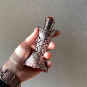 May include: An antique perfume bottle with an elaborate metal filigree design. The bottle has a brown-orange gemstone stopper and a red interior. The metalwork features floral patterns and openwork details.