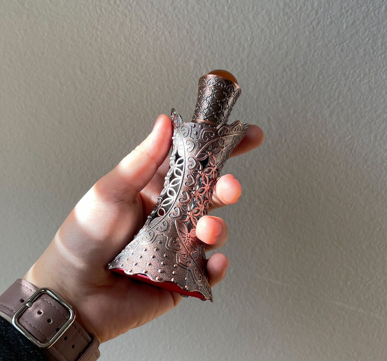 May include: An antique perfume bottle with an elaborate metal filigree design. The bottle has a brown-orange gemstone stopper and a red interior. The metalwork features floral patterns and openwork details.