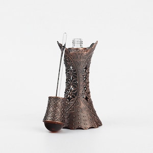 K&ouml;nnte beinhalten: Eine verzierte, bronzefarbene Parf&uuml;mflasche mit einem dekorativen Blumendesign. Die Flasche hat eine schlanke Glaspipette und eine kleine, runde, bernsteinfarbene Kugel. Das Design der Flasche ist aufwendig und detailliert.