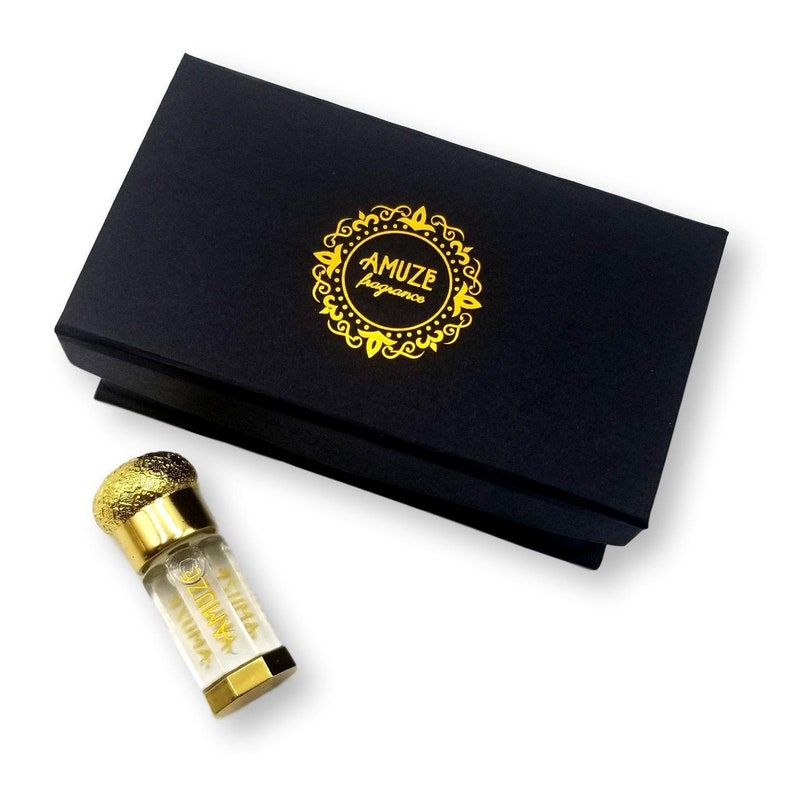 K&ouml;nnte beinhalten: Eine schwarze Schachtel mit einem goldenen Logo "AMUZE fragrance". Daneben eine kleine Parf&uuml;mflasche mit goldenen Akzenten und einem strukturierten goldenen Verschluss. Auf der Flasche steht "ZILINA AYURA".