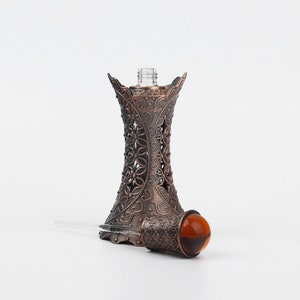 May include: An antique-style perfume bottle with a decorative bronze-coloured metal filigree holder. The bottle has a clear glass top and a brown glass stopper with a clear glass applicator. The filigree design features floral patterns.