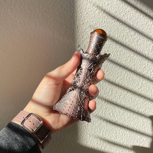 May include: An antique-style perfume bottle with a copper finish and intricate filigree detailing. The bottle has a decorative cap with a brown, gem-like stone. The bottle is held in a hand, with a pink watch strap visible in the foreground.