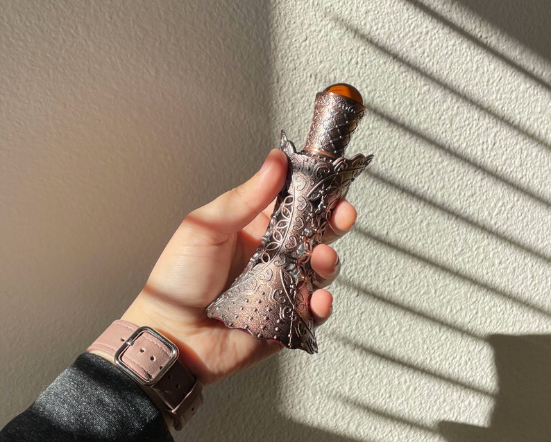 May include: An antique-style perfume bottle with a copper finish and intricate filigree detailing. The bottle has a decorative cap with a brown, gem-like stone. The bottle is held in a hand, with a pink watch strap visible in the foreground.