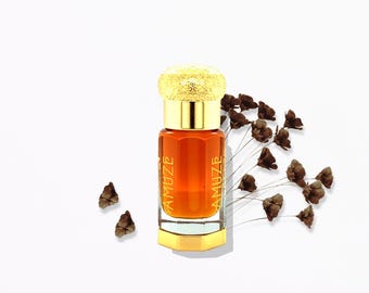 May include: A small, amber-colored perfume bottle with a gold cap and base. The bottle is labeled "AMUZE" on both sides. Dried brown flowers are arranged around the bottle on a white background. The cap has a textured design.