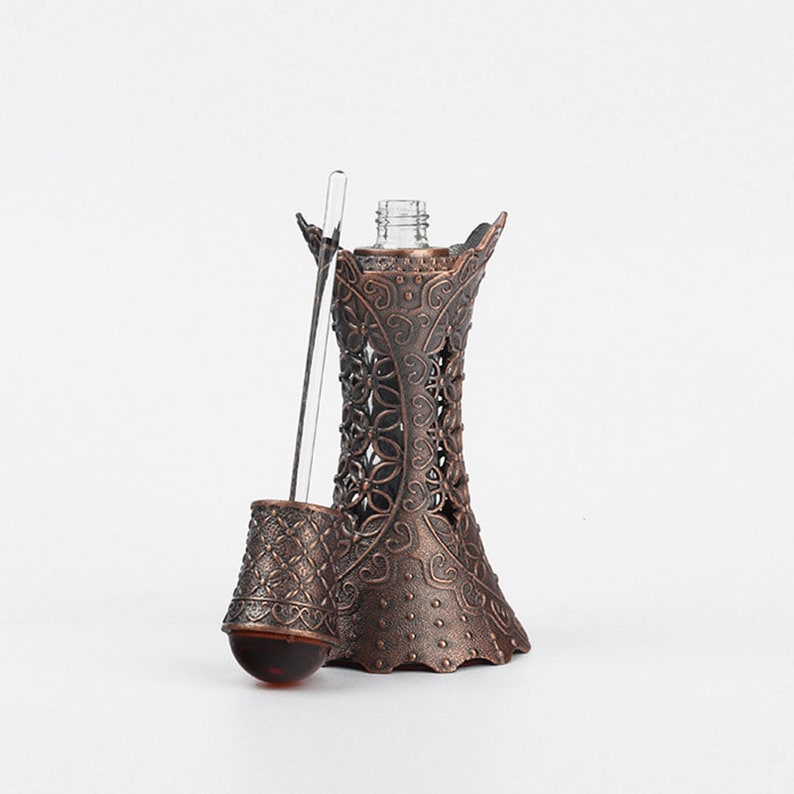 May include: An ornate, bronze-coloured perfume bottle with a decorative floral design. The bottle has a slender glass dropper and a small, round, amber-coloured bulb. The bottle's design is intricate and detailed.