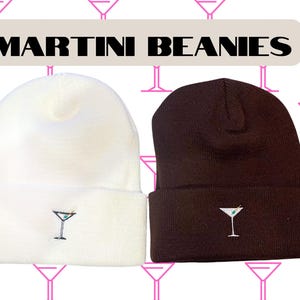 Op de afbeelding: Twee gebreide mutsen, een witte en een bruine, met een geborduurd martini-glas op de voorkant. De tekst "MARTINI BEANIES" wordt boven de mutsen weergegeven.