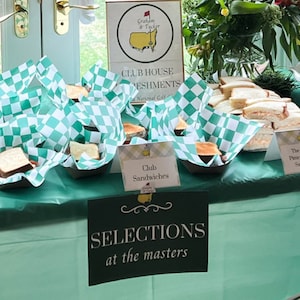 May include: A table covered in a light green tablecloth with a variety of sandwiches displayed on it. The sandwiches are wrapped in green and white checkered paper. There are signs that say "Club Sandwiches", "The Masters Pimento Cheese Sandwiches", and "On Par Pasta Salad".