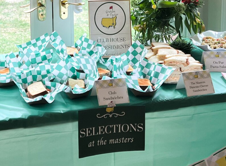 May include: A table covered in a light green tablecloth with a variety of sandwiches displayed on it. The sandwiches are wrapped in green and white checkered paper. There are signs that say "Club Sandwiches", "The Masters Pimento Cheese Sandwiches", and "On Par Pasta Salad".