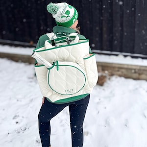 May include: A white quilted tennis bag with green trim and a circular pocket on the back. The bag has the text "HUT" in green on the front. The bag is being worn as a backpack by a person wearing a green and white knit hat.