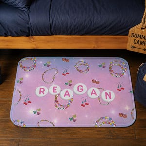 May include: A rectangular, pink and purple rug with the name "REAGAN" spelled out in white letters. The rug features a pattern of jewelry, butterflies, and earrings. A blue water bottle and a blue tote bag are visible.