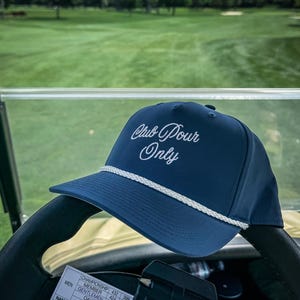 May include: A navy blue baseball cap with the text "Club Pour Only" in white script. The cap has a white rope detail along the brim. The cap is resting on a golf cart steering wheel, with a golf course in the background.
