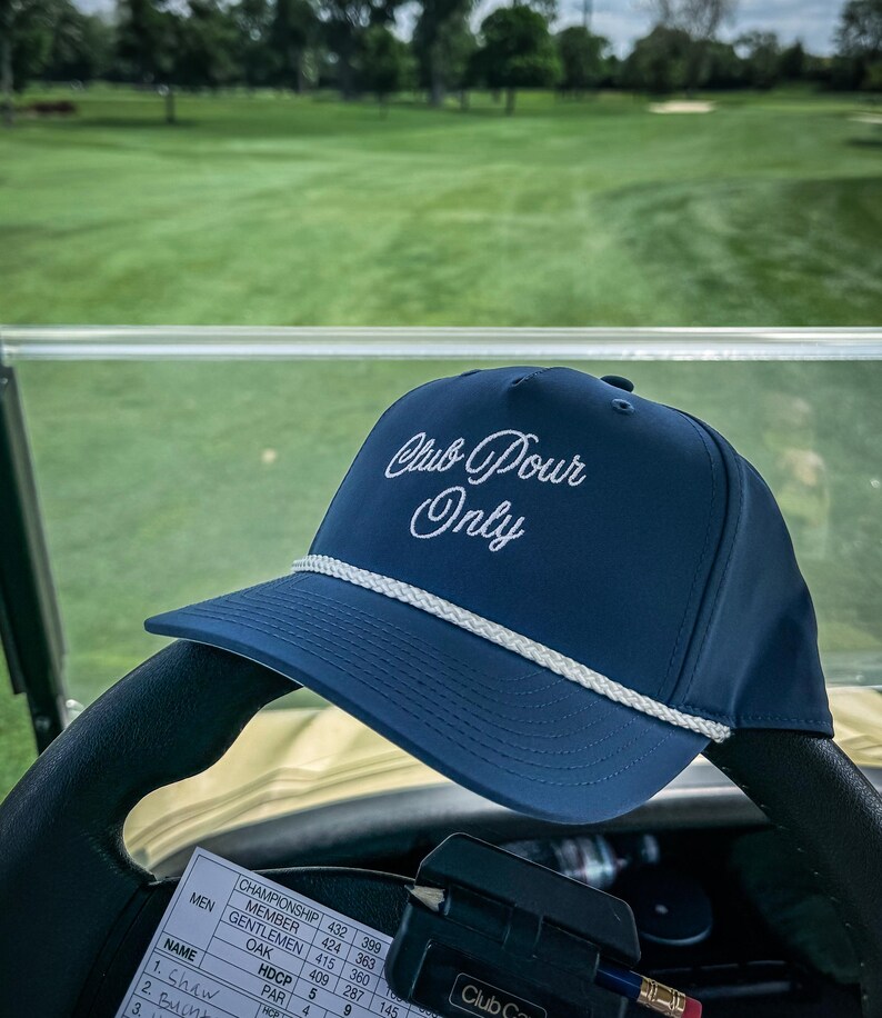 May include: A navy blue baseball cap with the text "Club Pour Only" in white script. The cap has a white rope detail along the brim. The cap is resting on a golf cart steering wheel, with a golf course in the background.