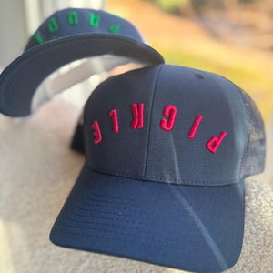May include: A navy blue baseball cap with a red embroidered word "PICKLE" on the front. The cap has a mesh back.