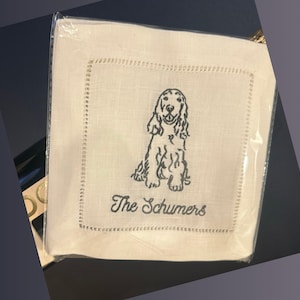 May include: Square white linen cocktail napkins with a decorative stitched border. Each napkin features a black embroidered Cocker Spaniel and the text "The Schumers". The napkins are packaged in clear plastic.