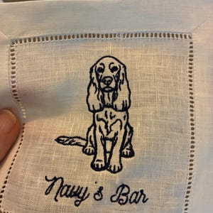 Könnte beinhalten: Weißes Cocktail-Servietten aus Leinen mit einem marineblauen Stickdesign. Das Design zeigt einen sitzenden Hund und die Worte "Navy's Bar" in Schreibschrift. Die Serviette hat einen dekorativen, genähten Rand.