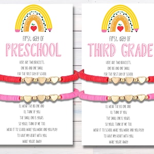 May include: Two bracelets, one pink and one red, with gold heart charms. The bracelets are on a white card with a rainbow and the text "First day of Preschool" and "First day of Third Grade".