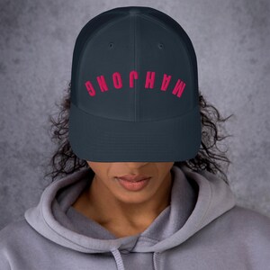 May include: A dark navy blue trucker hat featuring a mesh back and a solid front panel. The word "MAHJONG" is printed in a bold, bright pink font across the front, with the letters oriented in an upward-curving arc across the center of the cap.